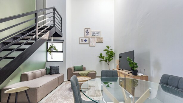 A modern living room with a glass dining table, grey sofas, a staircase, and framed artwork on the wall.