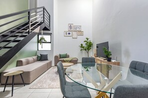 Una sala de estar moderna con una mesa de comedor de vidrio, sofás grises, una escalera y obras de arte enmarcadas en la pared.
