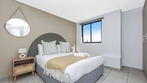 A modern bedroom with a large bed, a bedside table, a mirror, and a window with a view.