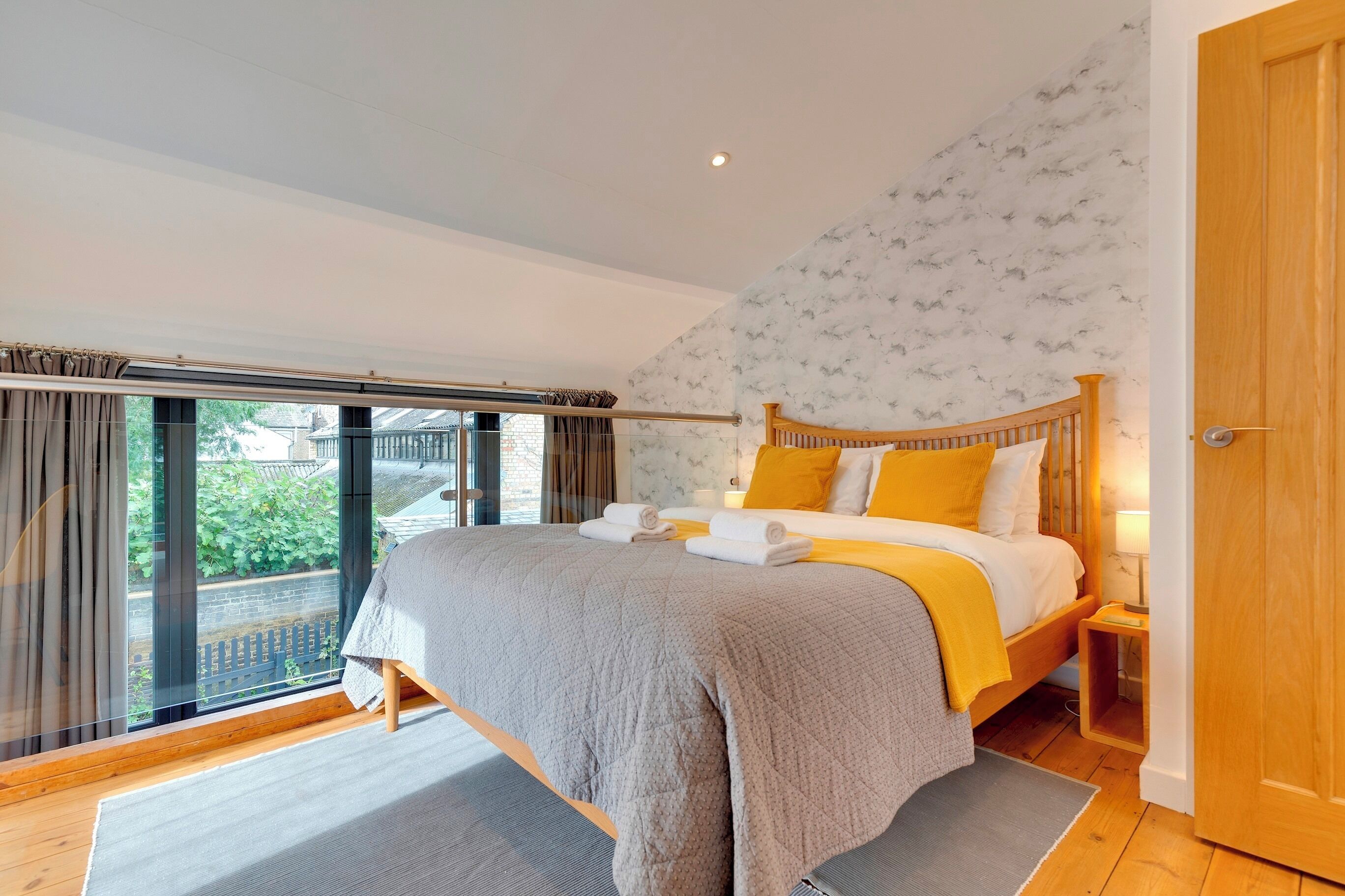 A bedroom with a large bed, a wooden headboard, a nightstand, and a view of greenery outside.