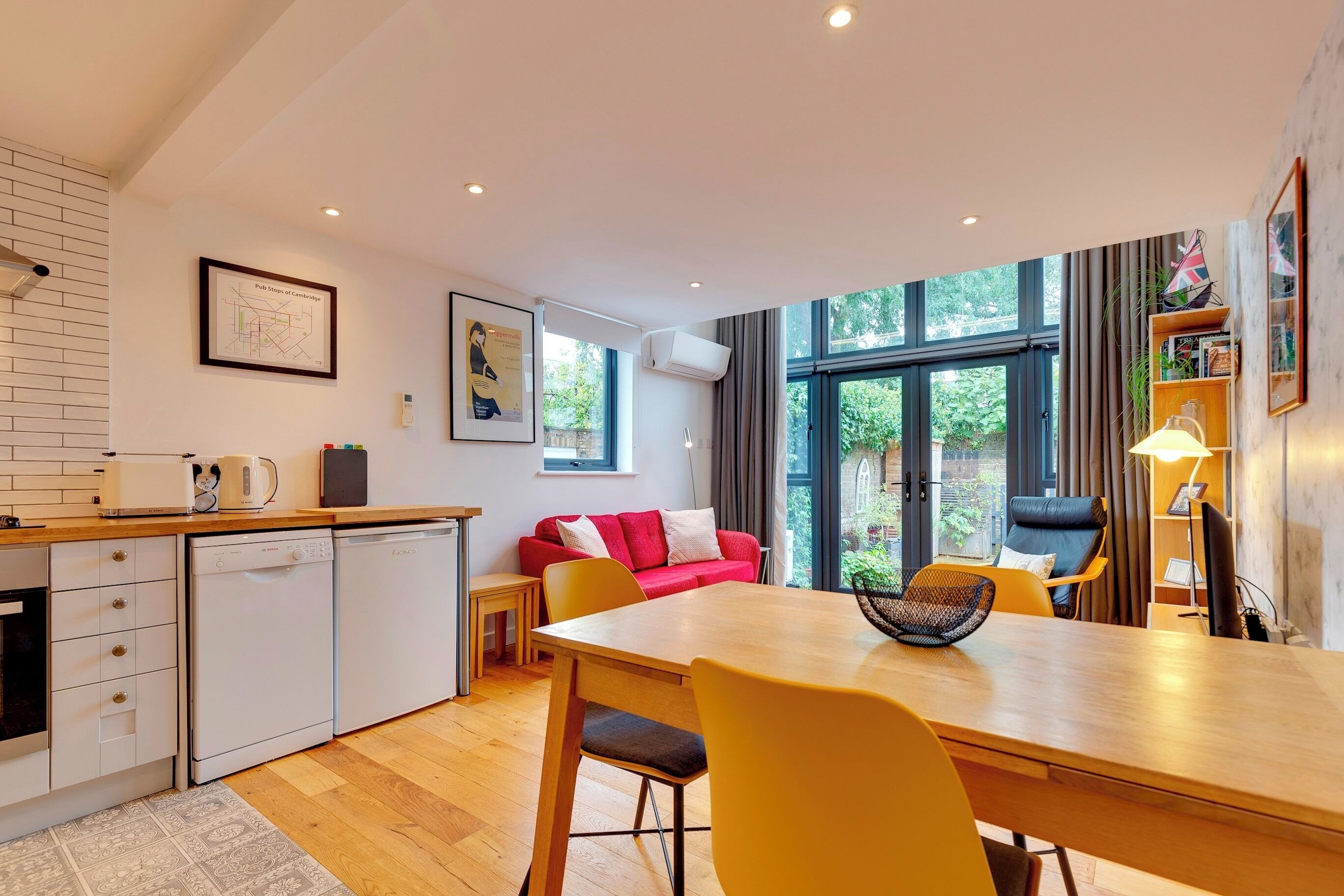 A modern kitchen with white appliances, a dining area with a wooden table and yellow chairs, a red sofa, and large windows offering a view of greenery.