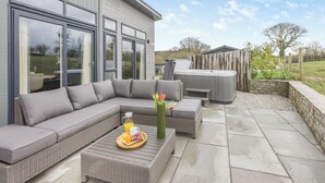 A modern outdoor living area with a sectional sofa, a hot tub, and a dining table set with a beverage and fruit. - 2 bedroom accommodation in Roadford Lake, near Launceston (Roadford Lake, near Launceston)