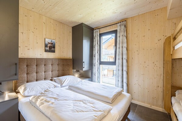 A bedroom with two beds, wooden walls, a large window with curtains, and a mounted picture of a cow.