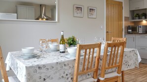 A dining area with a table set for four, including wine glasses, plates, and a bottle. There are wooden chairs and a white tablecloth with a leaf pattern. A potted plant and framed pictures are on the wall. - 3 bedroom accommodation in Arbroath (Arbroath)