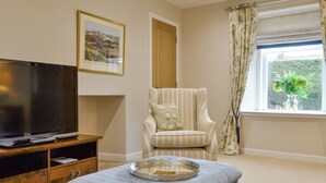 A living room with a television, a striped armchair, a small ottoman, and a window with curtains. - 3 bedroom accommodation in Arbroath (Arbroath)