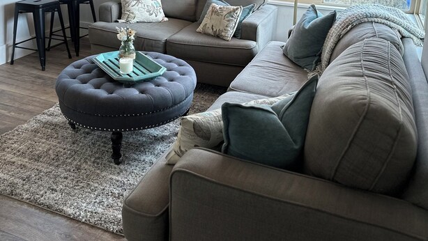 Un salon avec un canapé gris, un pouf rond et une petite table sur laquelle est posé un vase. Une fenêtre offre une vue sur l’extérieur.
