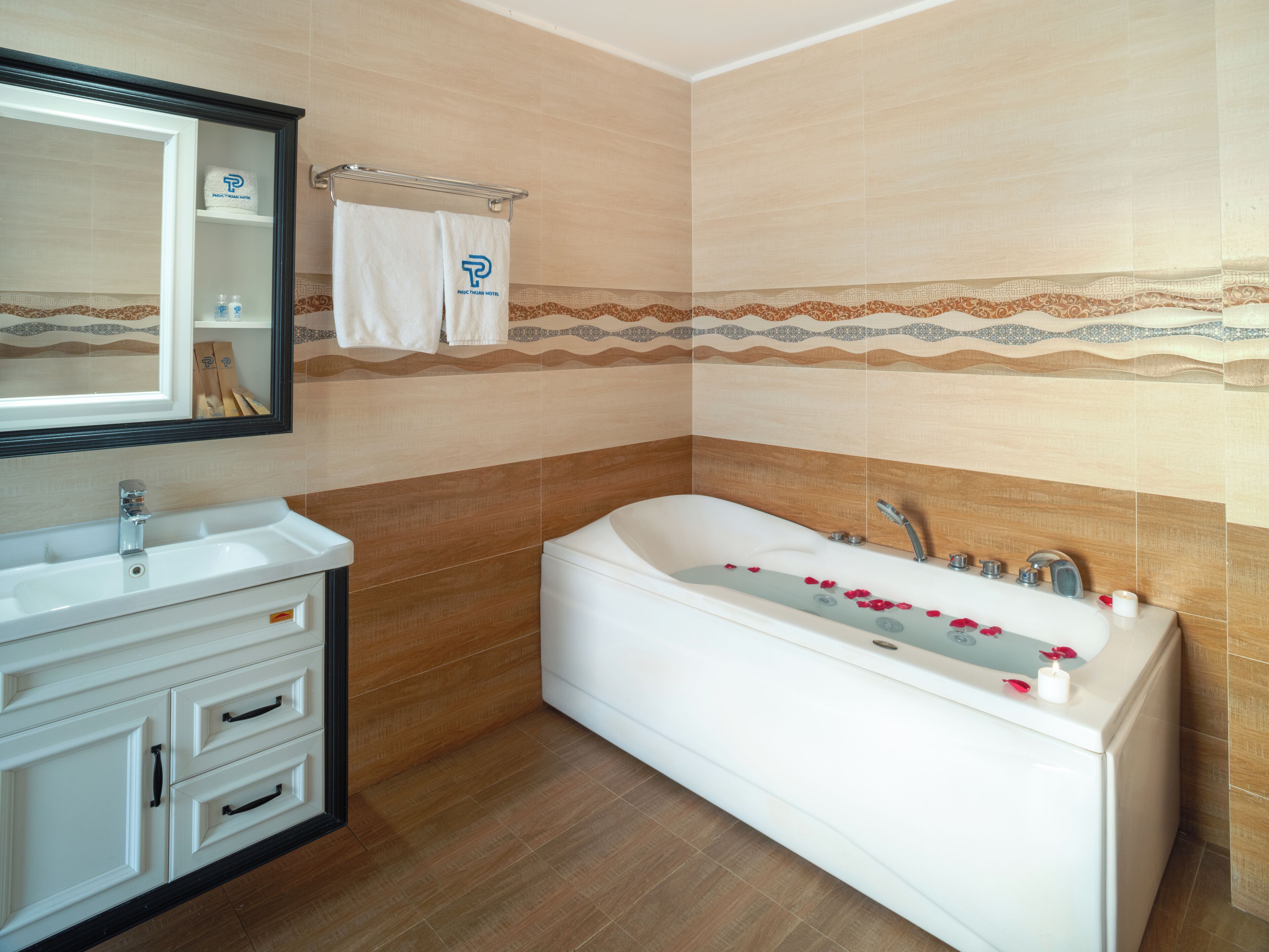 Un baño con una bañera blanca, un lavamanos y un espejo. Hay toallas colgadas en un perchero y una decoración de azulejos en la pared.