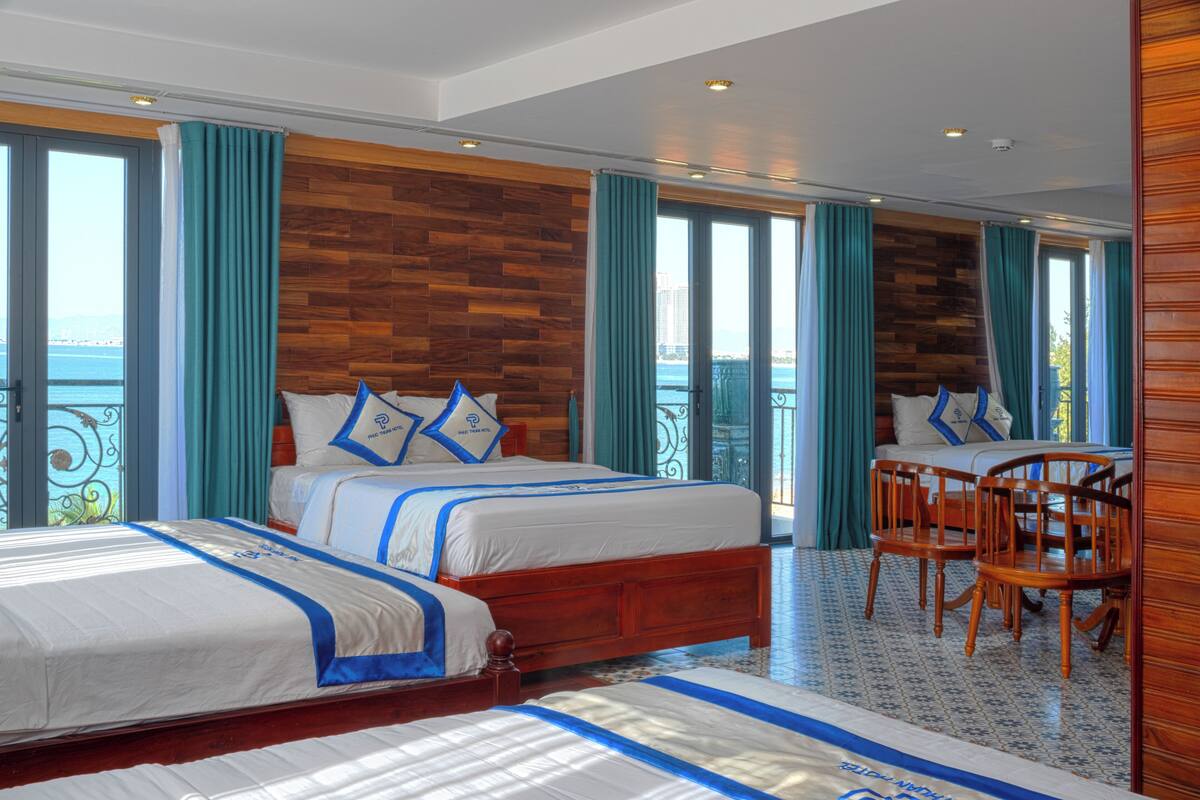 Une chambre d’hôtel avec deux lits, une tête de lit en bois, un balcon avec vue et un coin repas avec des chaises.