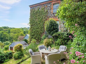 A stone building with ivy, surrounded by lush greenery and a patio with wicker furniture.