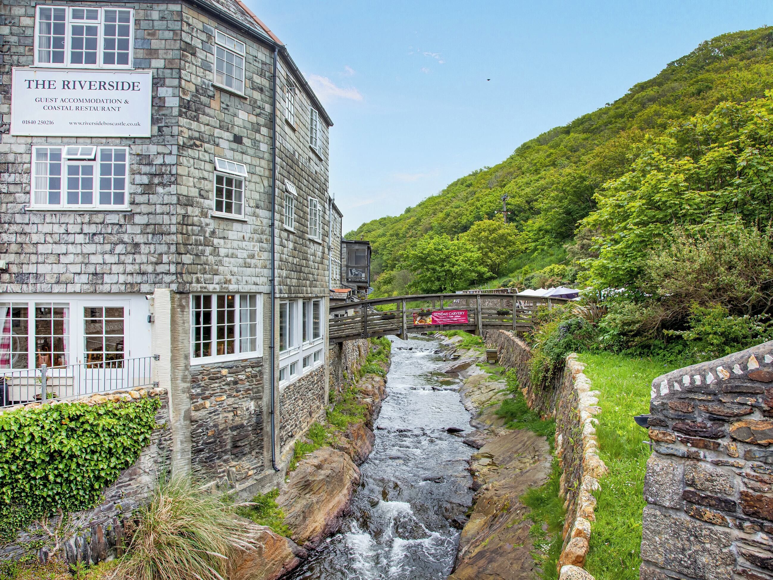 A stone building labeled "The Riverside" with a signboard, a small bridge, and a flowing stream.