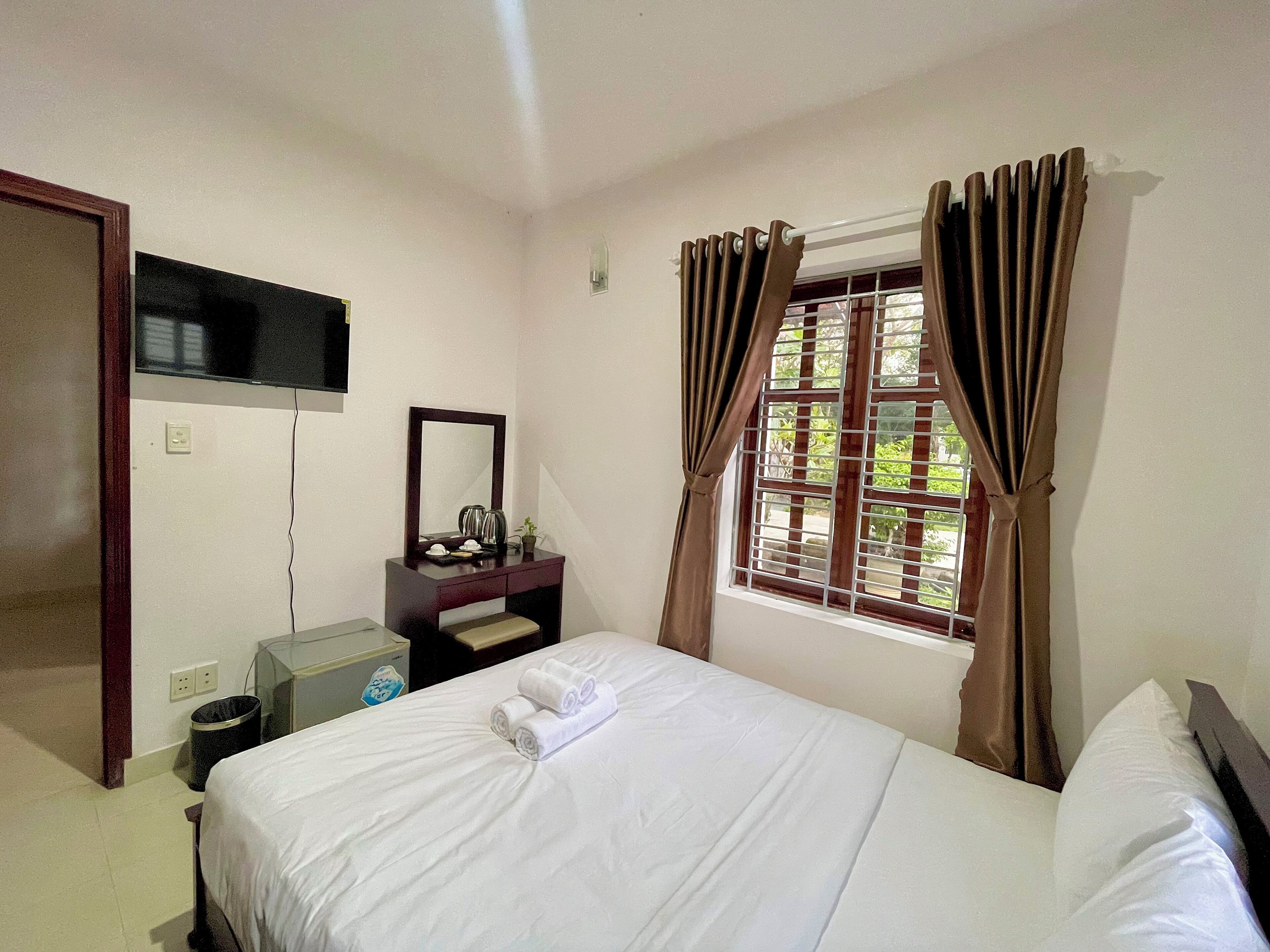 Quarto de hotel com cama, mesa de cabeceira com espelho, pequena escrivaninha e janela com cortinas.