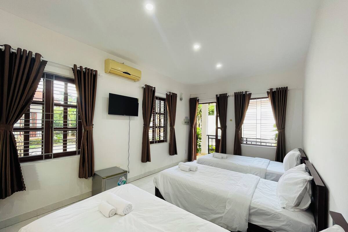 A hotel room with two beds, a flat-screen TV, an air conditioning unit, and windows with curtains.