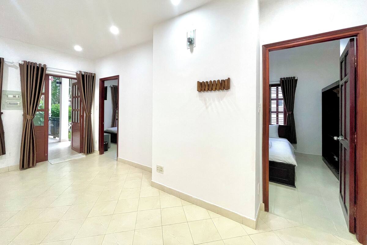 A hotel room with a bed, a small table, and a chair. There is a bathroom with a toilet and sink. The room has a tiled floor and white walls.