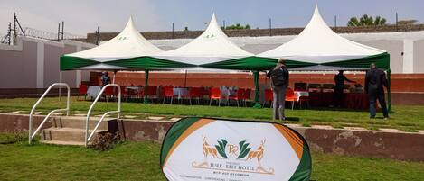 A tented outdoor event with tables and chairs, a sign for the Turk-Reef Hotel, and a grassy area with steps.