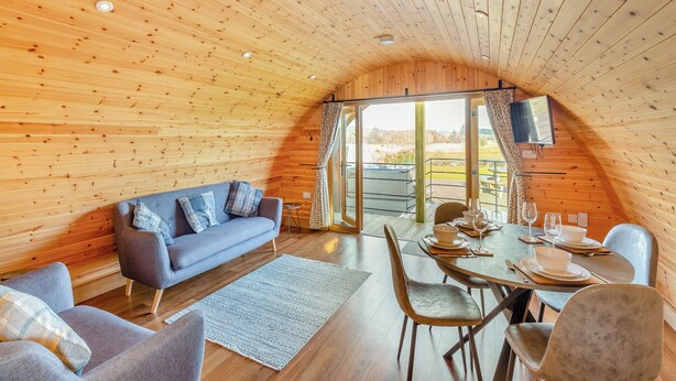 L’intérieur chaleureux d’un chalet avec un plafond en bois, un canapé, une table à manger dressée pour deux personnes et une grande fenêtre donnant sur le paysage extérieur.