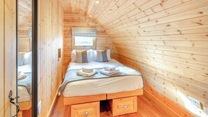 Une pièce chaleureuse aux murs en bois, avec un lit, une fenêtre agrémentée d’une lambrequin et un luminaire au plafond.
