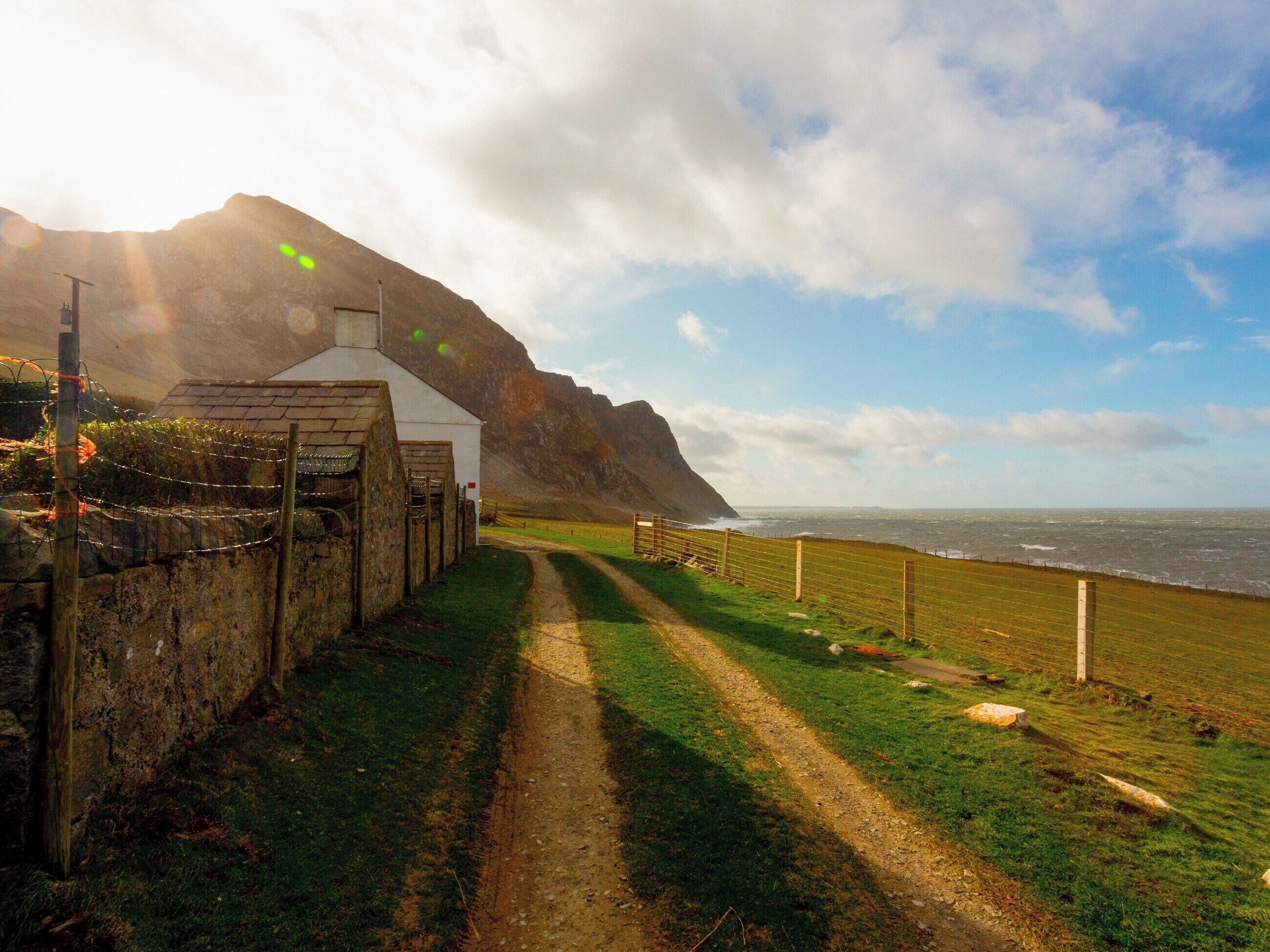 A coastal path leading to a white building with a stone wall and a fenced area.