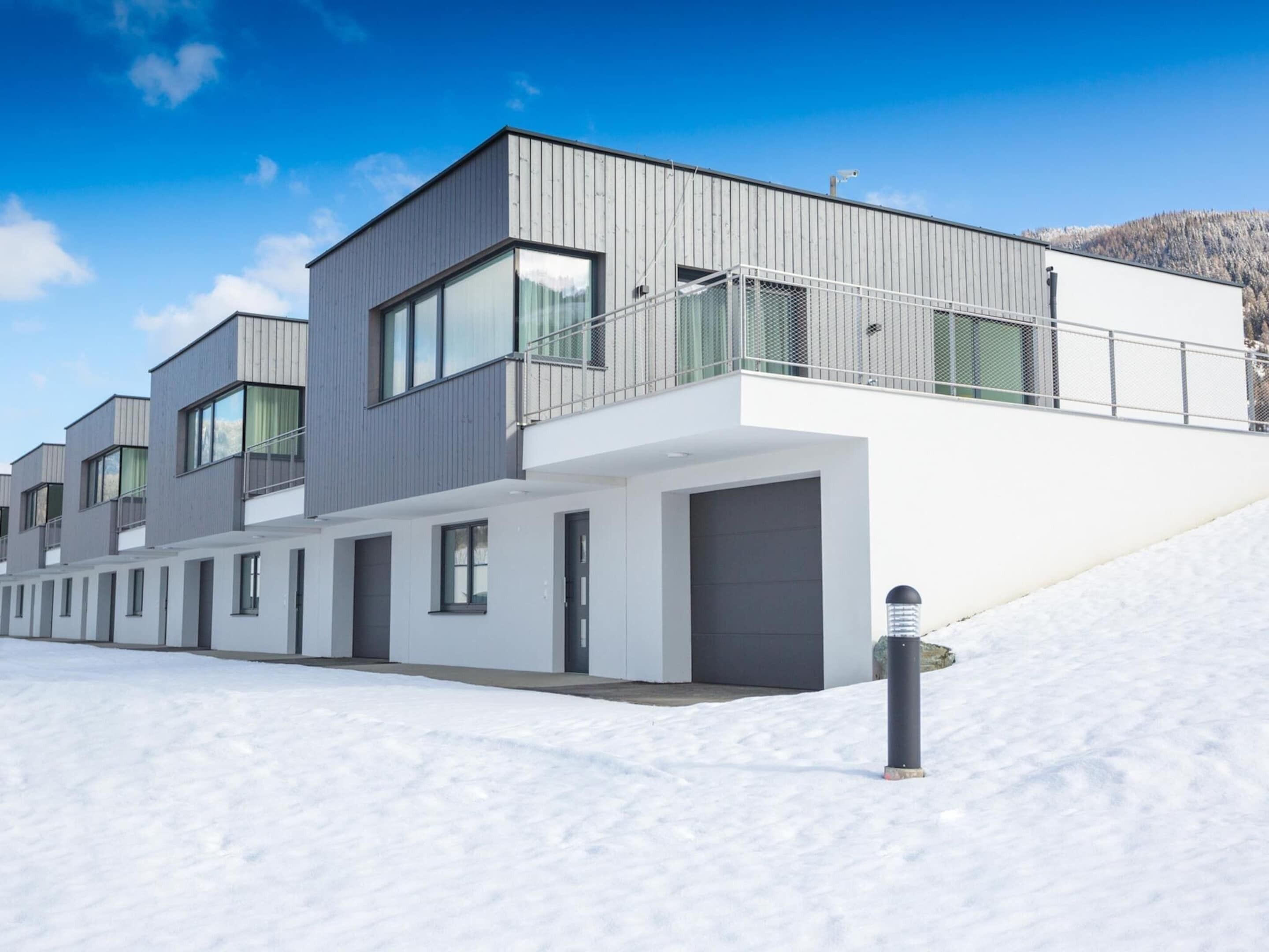 A modern multi-story residential building with large windows and a flat roof, situated on a snowy slope.