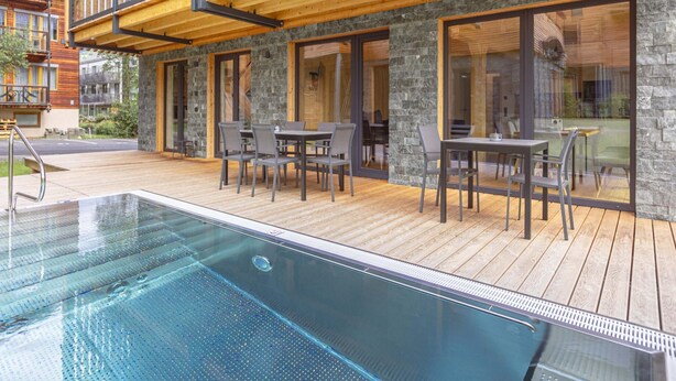 A modern outdoor pool area with wooden decking, stone walls, and glass doors leading to a dining area with outdoor seating. - Spacious Apartment With Pool (Sankt Georgen am Kreischberg)