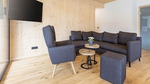 A modern living room with a sectional sofa, a small round table with a plant, and a flat-screen TV mounted on a wooden wall. - Spacious Apartment With Pool (Sankt Georgen am Kreischberg)