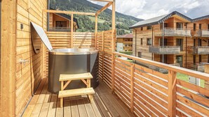 Una terraza de madera con un jacuzzi y escalones de madera, con vistas a un paisaje montañoso y otras construcciones de madera.