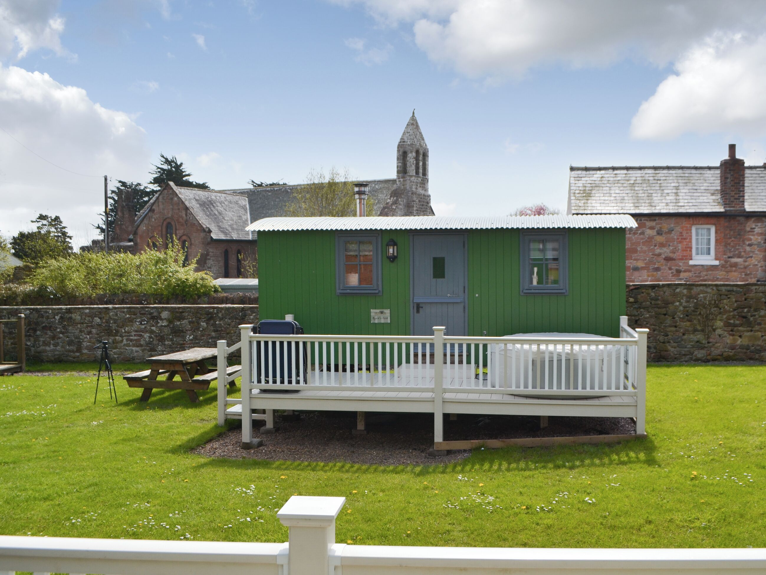 A small green cottage with a white porch, a stone church in the background, and a picnic table.