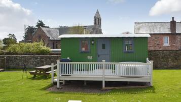 A small green cottage with a white porch, a stone church in the background, and a picnic table.