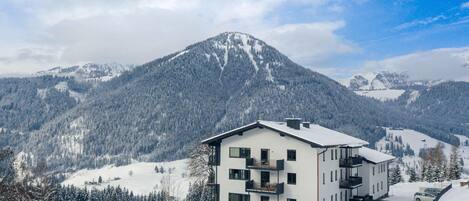 A modern multi-story building with balconies, surrounded by snow-covered mountains and trees.