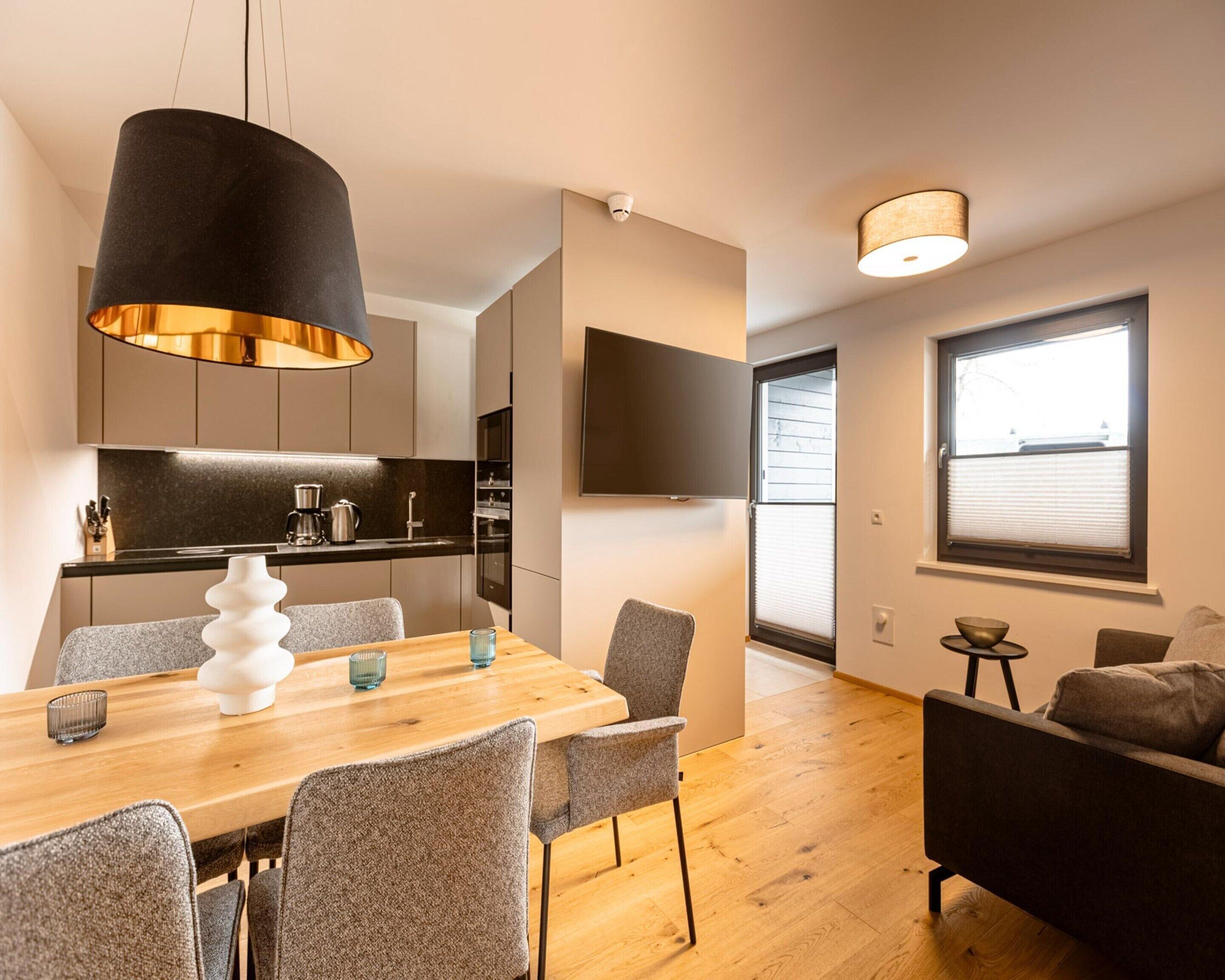 Une cuisine moderne avec une table et des chaises en bois, un luminaire suspendu noir et un téléviseur à écran plat fixé au mur.