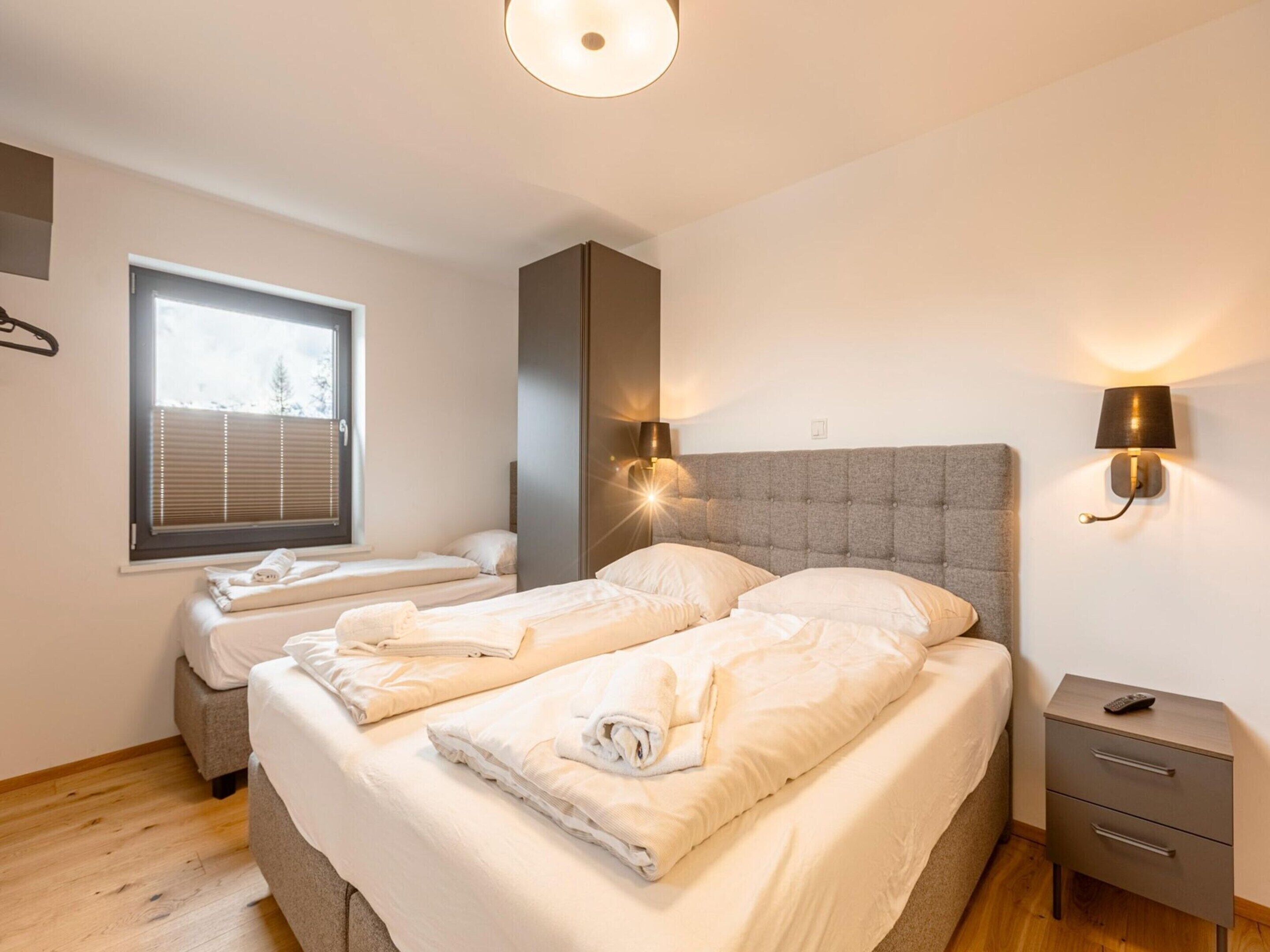 A bedroom with two beds, a bedside table, a nightstand, a window with blinds, and wall-mounted lights.