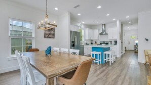 A modern kitchen with white cabinets, a central island, and a dining area with a wooden table and white chairs. - Seabbatical: Private heated pool, Free golf cart, 2min to beach, Game room (Miramar Beach)