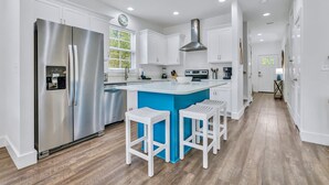 A modern kitchen with white cabinets, a stainless steel refrigerator, a blue island with bar stools, and a window with blinds. - Seabbatical: Private heated pool, Free golf cart, 2min to beach, Game room (Miramar Beach)