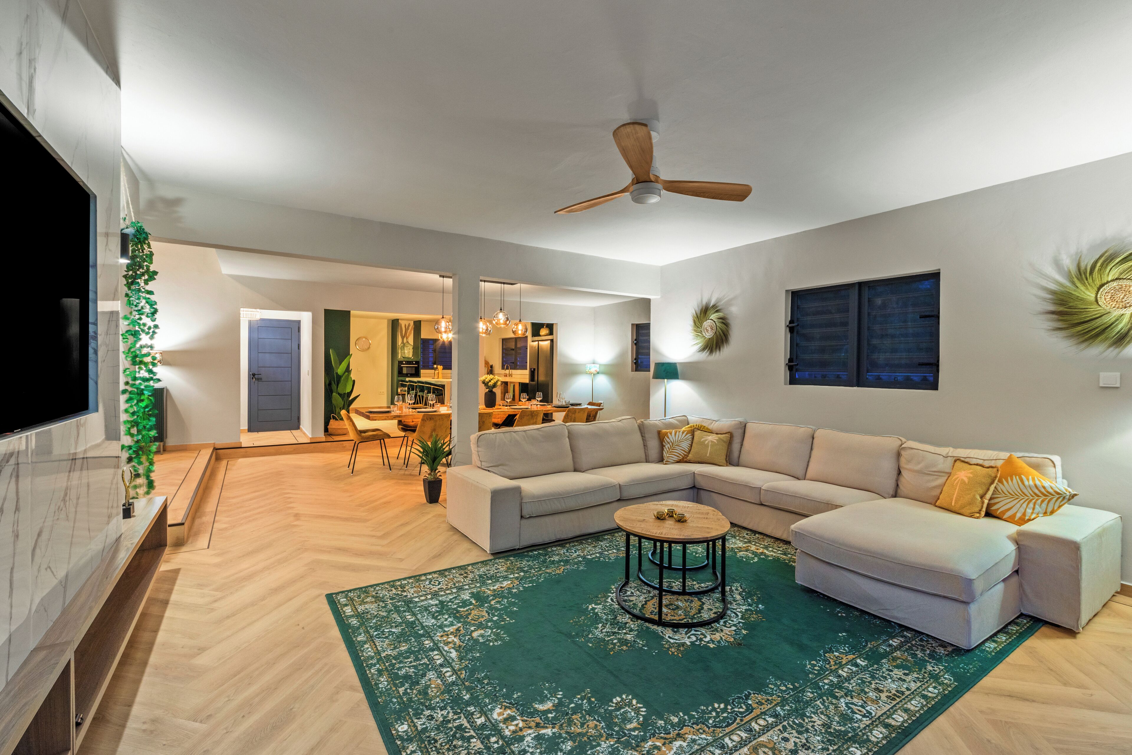A spacious living room with a large sectional sofa, a round coffee table, and a ceiling fan. A TV is mounted on the wall, and there is a dining area visible in the background.