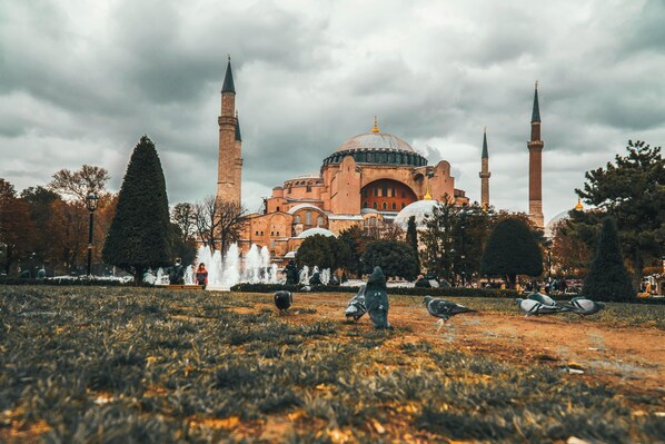 Un bâtiment historique doté de plusieurs minarets et d’un grand dôme central, entouré d’un parc avec une fontaine et des pigeons.