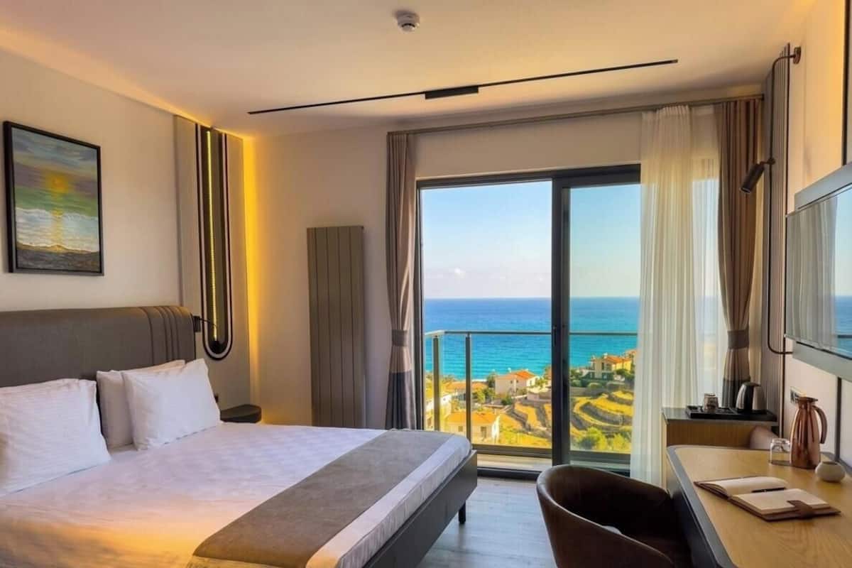 Ein Hotelzimmer mit einem Bett, einem Schreibtisch, einem Stuhl, einem großen Fenster mit Meerblick und einem Gemälde an der Wand.