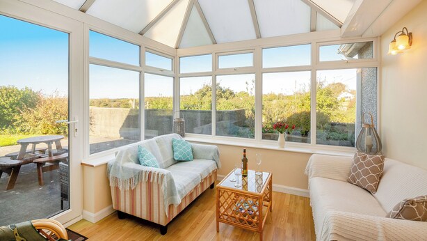 A conservatory with a glass roof, a sofa, a table with a bottle and glass, and a view of greenery outside.