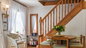 Ein gemütliches Zimmer mit einer Holztreppe, einem Korbstuhl, einem runden Holztisch mit Stühlen und einem Bücherregal.