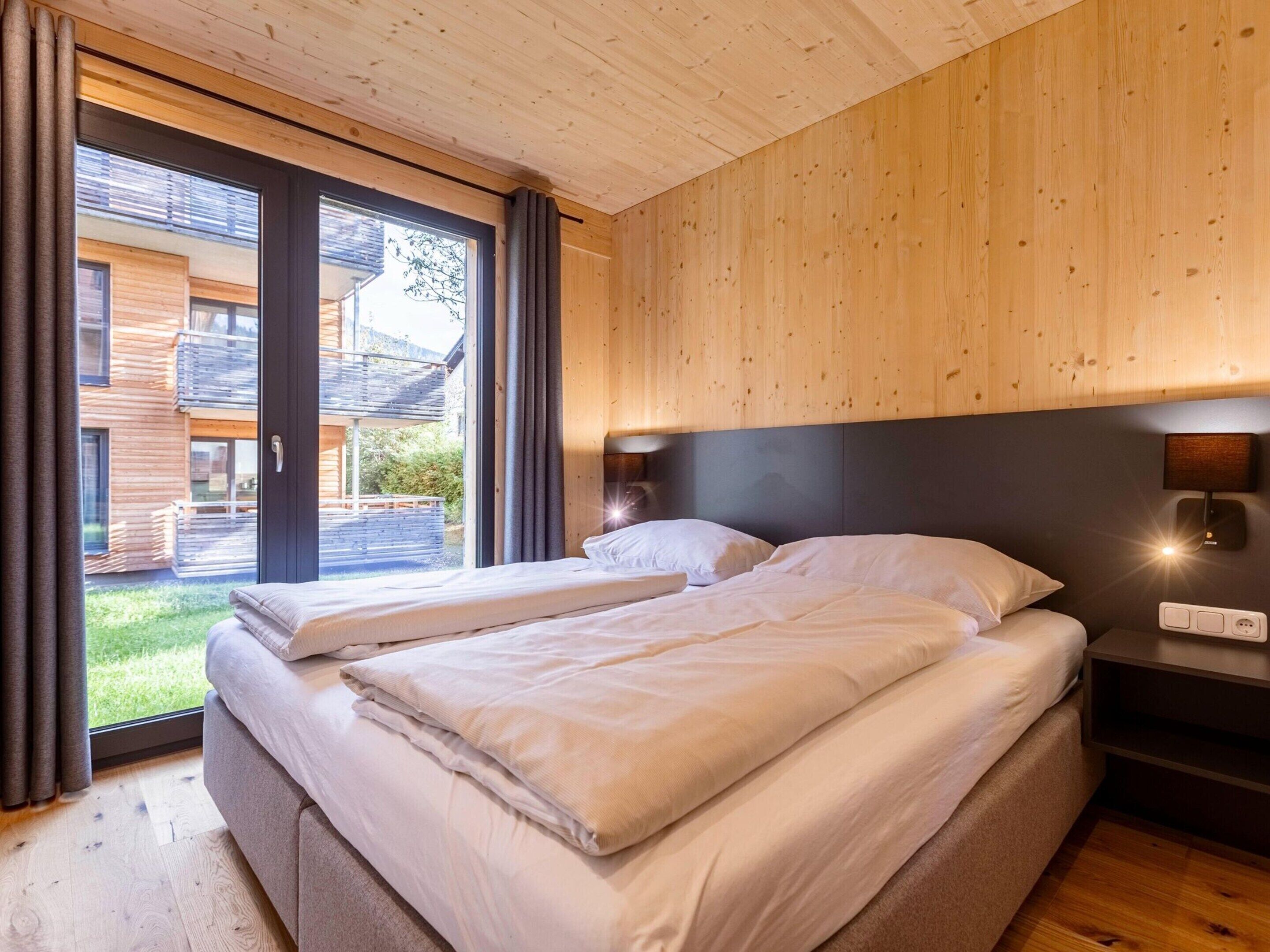 A bedroom with two beds, wooden walls, a large window, and a view of a building outside.