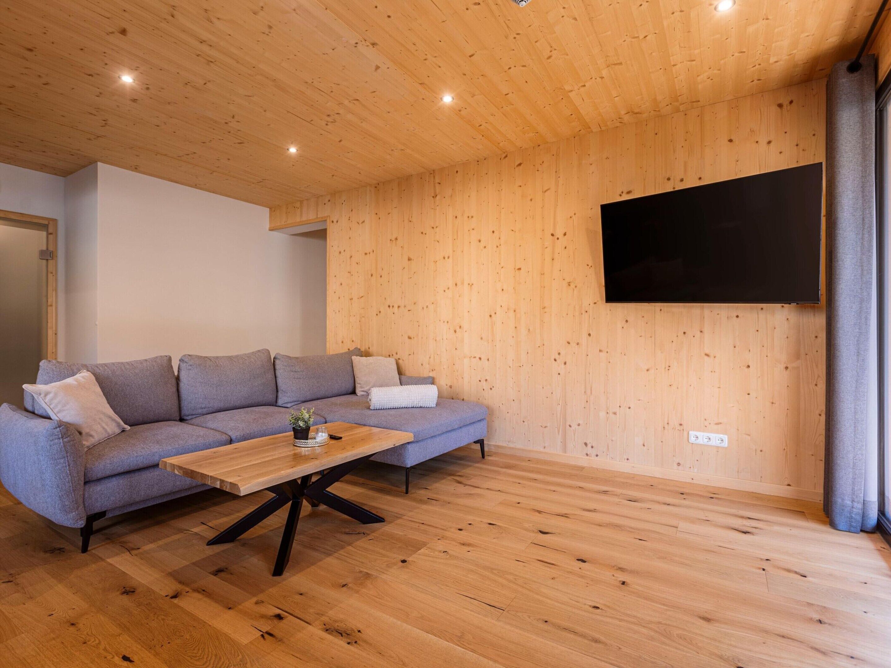 Un salon moderne avec un plafond en bois, un canapé gris, une table basse en bois et un téléviseur à écran plat fixé au mur.