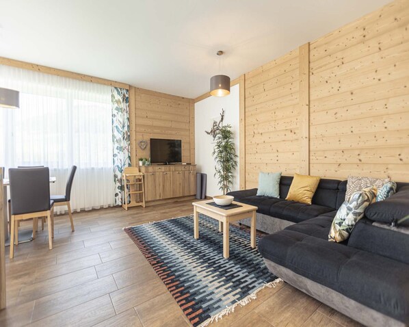 A modern living room with a wooden floor, a black sofa, a wooden coffee table, a TV on a wooden cabinet, and a dining area with a table and chairs.