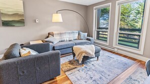 A modern living room with a grey sofa, a side table, a floor lamp, and large windows with a view of greenery. - Super Cozy Studio Near Slopes And Shuttle Bus WiFi (Snowshoe)