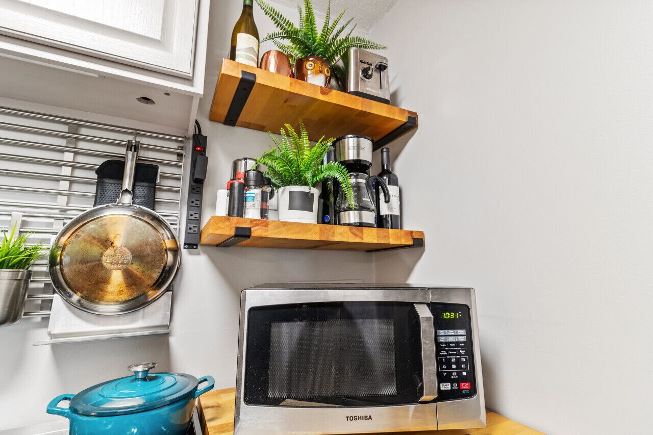 A kitchen with a microwave, a pot, and a shelf with various items including a plant and a bottle.