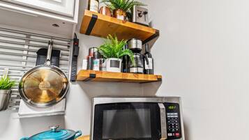 A kitchen with a microwave, a pot, and a shelf with various items including a plant and a bottle.