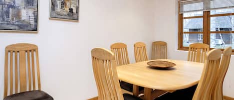A dining area with a wooden table and chairs, framed artwork on the wall, and a window with blinds.