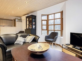 Una sala de estar moderna con un sofá de cuero, un sillón gris, una mesa de comedor de madera y un ventanal con vistas a árboles.