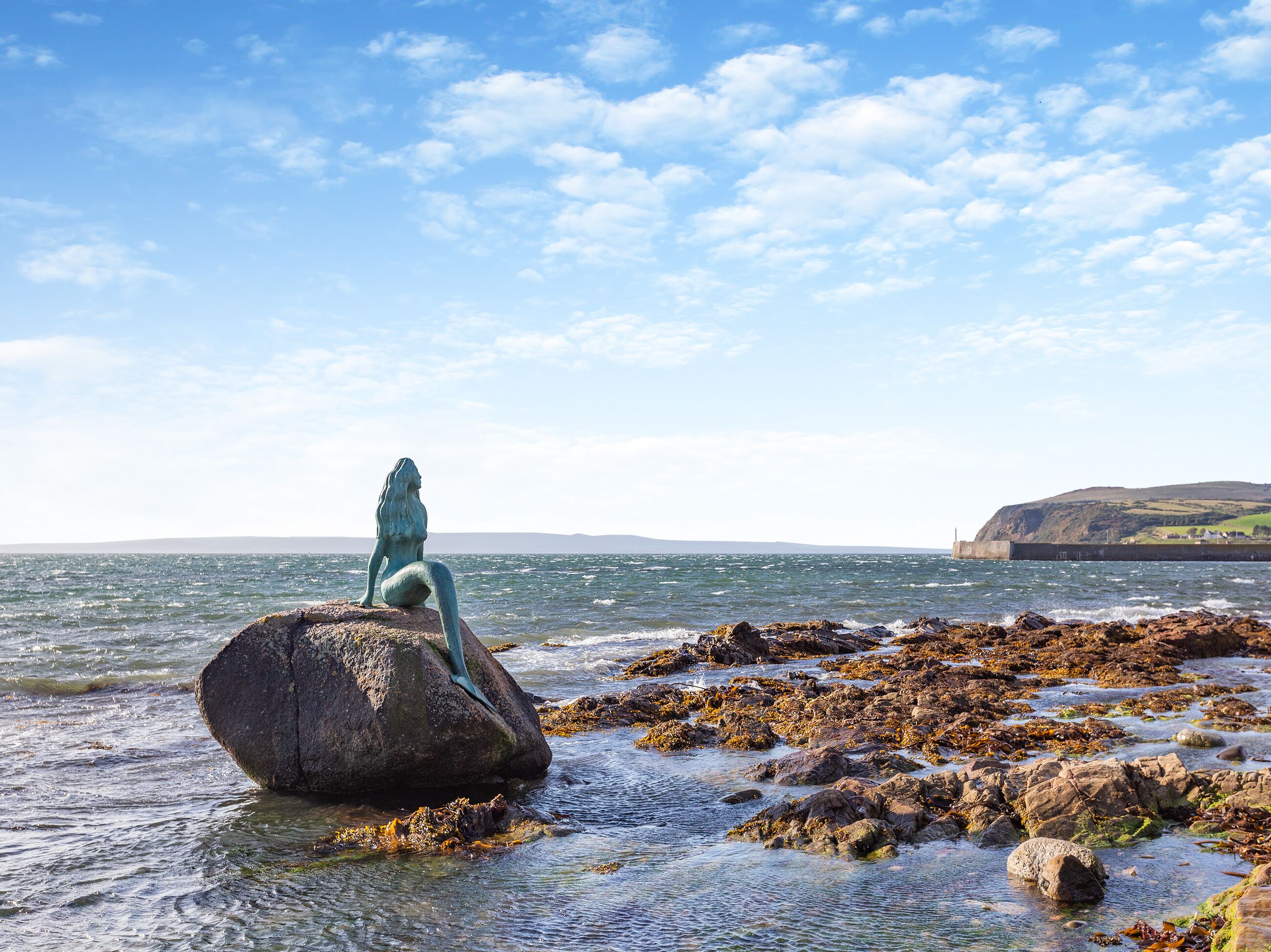 A mermaid statue on a rocky shore with a view of the sea and distant hills.