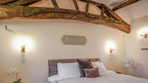 A bedroom with a wooden ceiling, a bed with white and brown bedding, a bedside table with a plant, and a 'Be our guest' sign on the wall. - 1 bedroom accommodation in Combs, near Chapel-en-le-Frith (Combs, near Chapel-en-le-Frith)