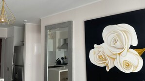 Uma sala de estar moderna com um sofá cinza, uma pintura de rosa preta e branca, e uma cozinha visível através de uma porta aberta.