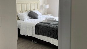 Cama bem arrumada com cabeceira, luminária ao lado e toalha dobrada sobre piso de madeira.