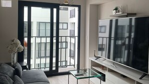 Uma sala de estar moderna com um sofá preto, uma mesa de centro de vidro, uma televisão de tela plana e uma varanda com vista para edifícios.